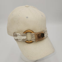Load image into Gallery viewer, An off-white ribbed knit baseball cap with a decorative woven band featuring a large gold-tone ring and a small brown button, displayed on a white mannequin head against a light gray background.