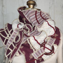 Load image into Gallery viewer, A chunky knit scarf with a large bow, featuring burgundy and cream striped panels, textured cream knit sections, and a brown wooden ring accent, displayed on a cream-colored mannequin torso against a white wooden wall.