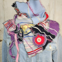 Load image into Gallery viewer, A handmade patchwork knit scarf displayed on a denim jacket mannequin against a light wood paneled background. The scarf features a mix of colorful knit fabrics including light blue, purple, pink, black, and white, with a prominent red button accent.