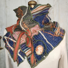 Load image into Gallery viewer, A handmade, multi-layered knit scarf or cowl displayed on a white ribbed sweater dress form. The scarf features a complex patchwork design with blue, green, pink, and orange knit fabrics, striped bands, frayed edges, and a decorative cream-colored brooch.