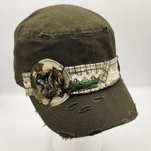 Load image into Gallery viewer, A distressed olive green cap with a tattered brim, featuring a decorative band with a camouflage flower embellishment and a green landscape print, 
