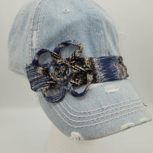 Load image into Gallery viewer, A light blue distressed denim baseball cap with a decorative dark blue and black patterned fabric flower accessory on the front, displayed on a white mannequin head against a light gray background.
