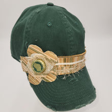 Load image into Gallery viewer, A dark green distressed baseball cap featuring a decorative woven band with a flower applique and a spotted cow bottle cap embellishment on the front.
