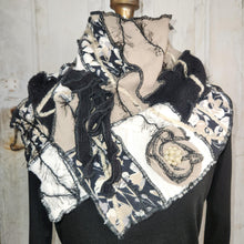 Load image into Gallery viewer, A handmade patchwork scarf displayed on a black mannequin torso against a white wooden wall. The scarf features a mix of black and beige fabrics, including floral patterns, lace, frayed edges, and a central patch with a pearl-like flower.
