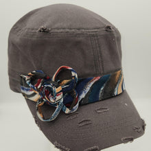 Load image into Gallery viewer, A distressed dark gray fabric cap with a colorful patterned fabric flower on the brim, displayed on a white mannequin head against a light gray background.
