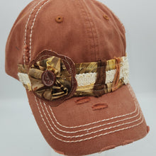 Load image into Gallery viewer, A distressed rusty orange baseball cap with white contrast stitching, featuring a decorative fabric band with camouflage, lace, and a fabric flower embellishment, displayed on a white mannequin head against a light gray background.
