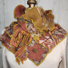 Load image into Gallery viewer, A handmade, highly textured ruffled cowl scarf displayed on a cream-colored ribbed sweater dress form. The scarf features a complex patchwork design with mustard yellow chunky knit sections, rust-red floral appliques, and a patterned fabric base in brown, beige, and gray, finished with a metallic gold-tone button accent.

