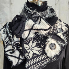 Load image into Gallery viewer, A black and white knit scarf with a Nordic snowflake pattern, draped on a mannequin. The scarf features a large black button, a knotted black cord detail, and a frayed, textured edge, styled over a black top against a rustic white wooden background.
